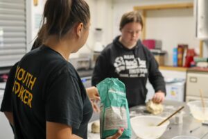 Catering course, a youth worker in the foreground holding a bag of flour, while the background shows a bowl on a table with young person, out of focus kneading dough