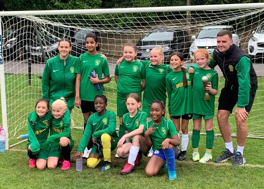 The Hunslet Club Girls Football under 10s team posing together in front of a football goal. All wearing matching green Hunslet Club uniforms. 