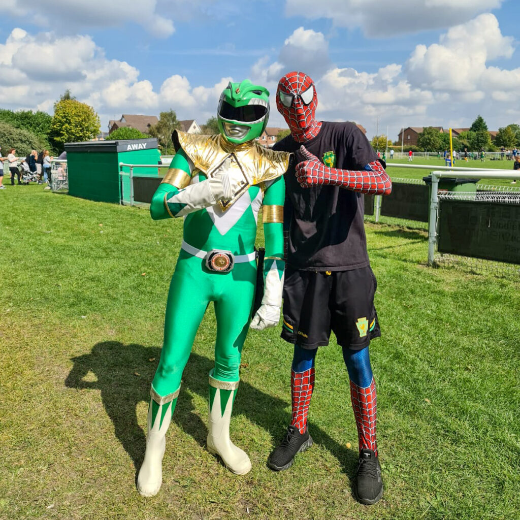 Two people dressed up one as the Green Power Ranger, the other as Spiderman. Posing together on a field with thumbs up.