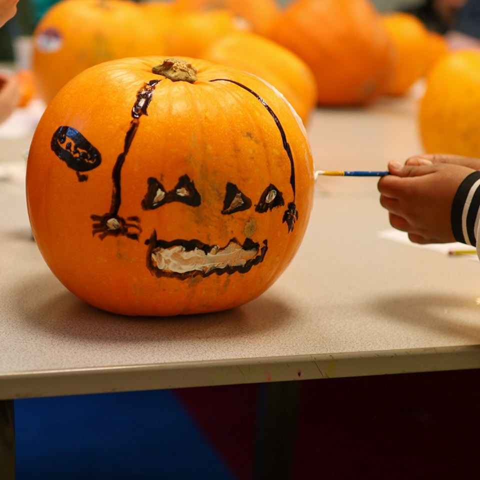 Painting a pumpkin. The pumpkin has a spider painted on the side, along with a face. Someone is still painting the pumpkin, as a hand can be seen on the right. 