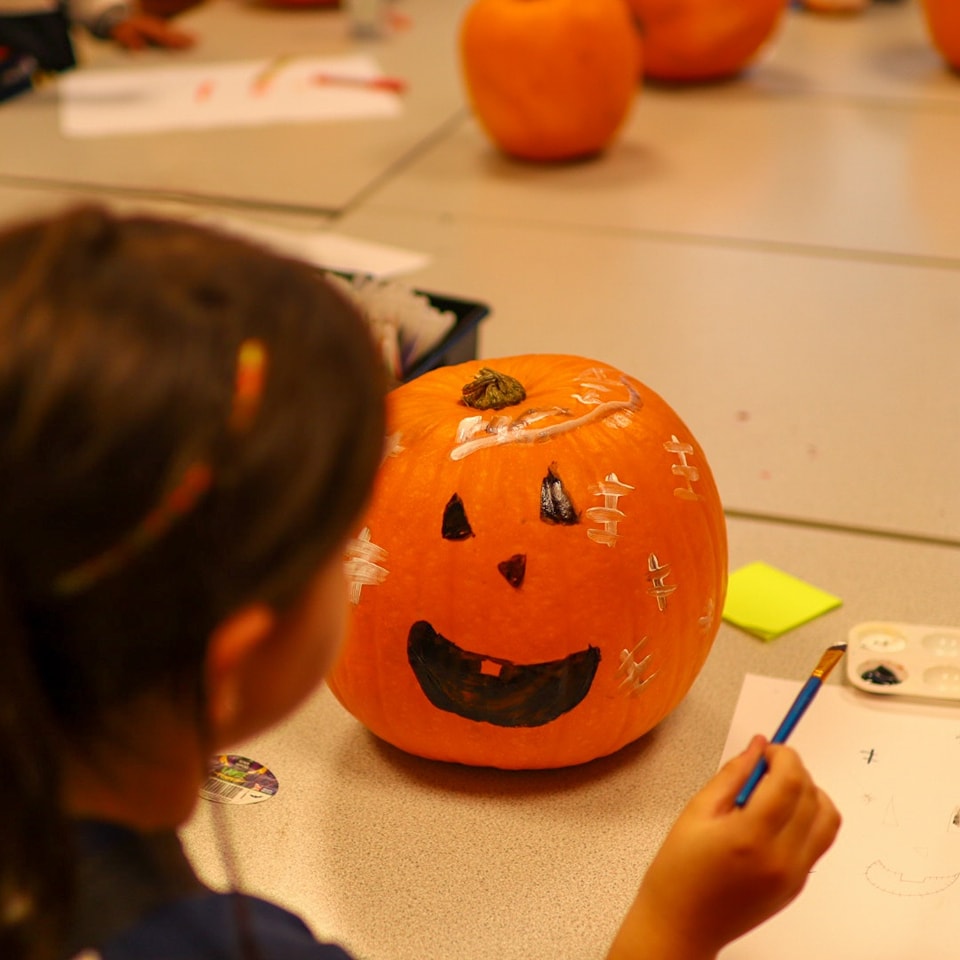 A young person painting a pumpkin. The pumpkin has a smiling face, while a young person holds a paint brush, ready to paint. 