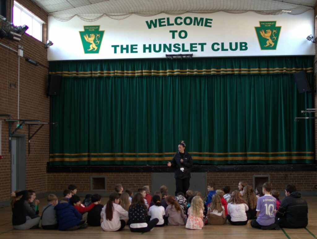 A group of young people sat on the floor of a hall at The Hunslet Club, with a West Yorkshire Police Officer standing at the front, talking about firework safety. 