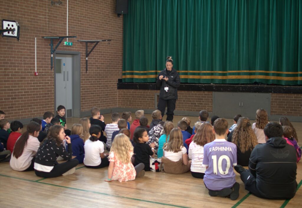A group of young people sat on the floor of a hall, with a West Yorkshire Police Officer standing at the front, talking about firework safety. 