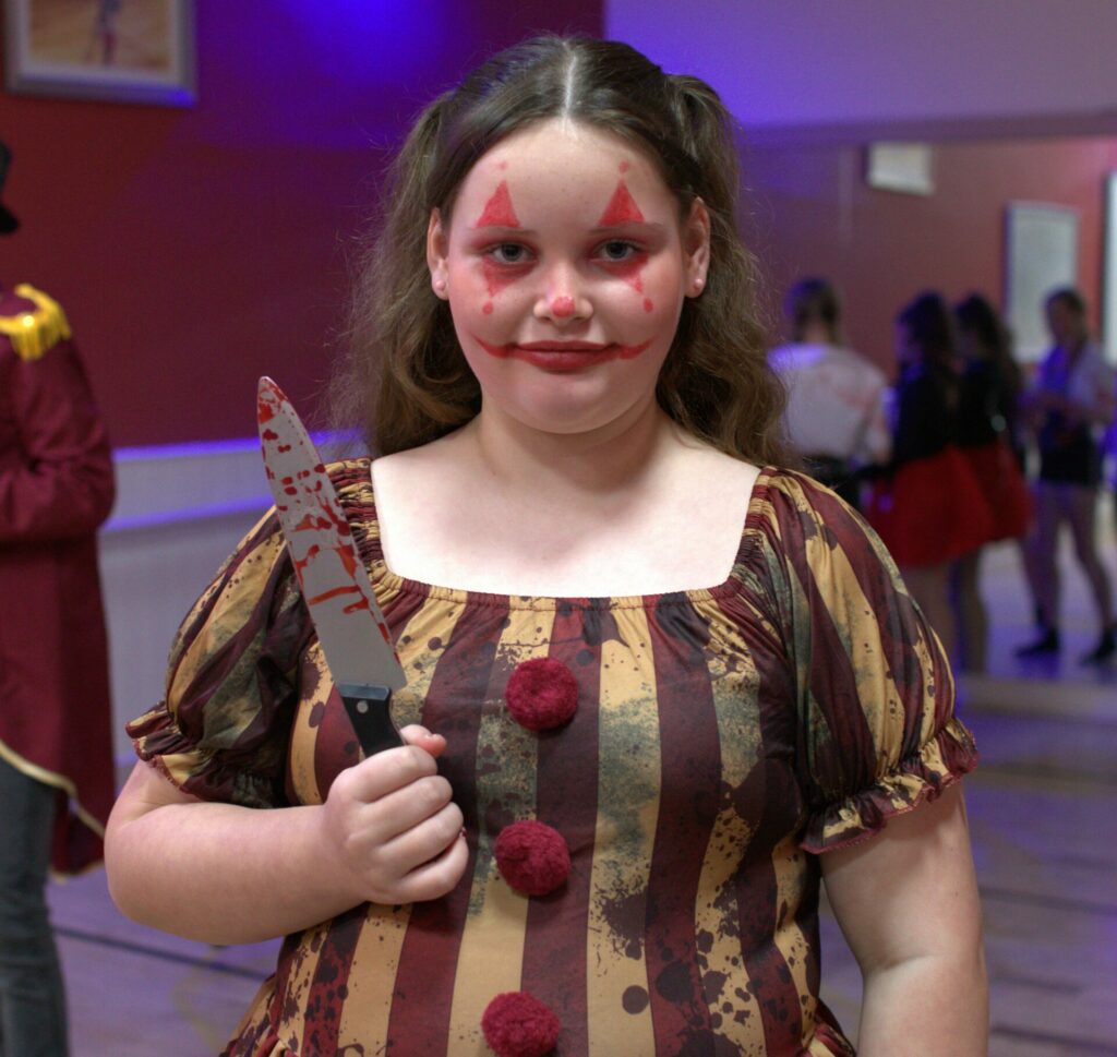 A girl dressed as a scary clown, holding a fake knife, covered in blood. 