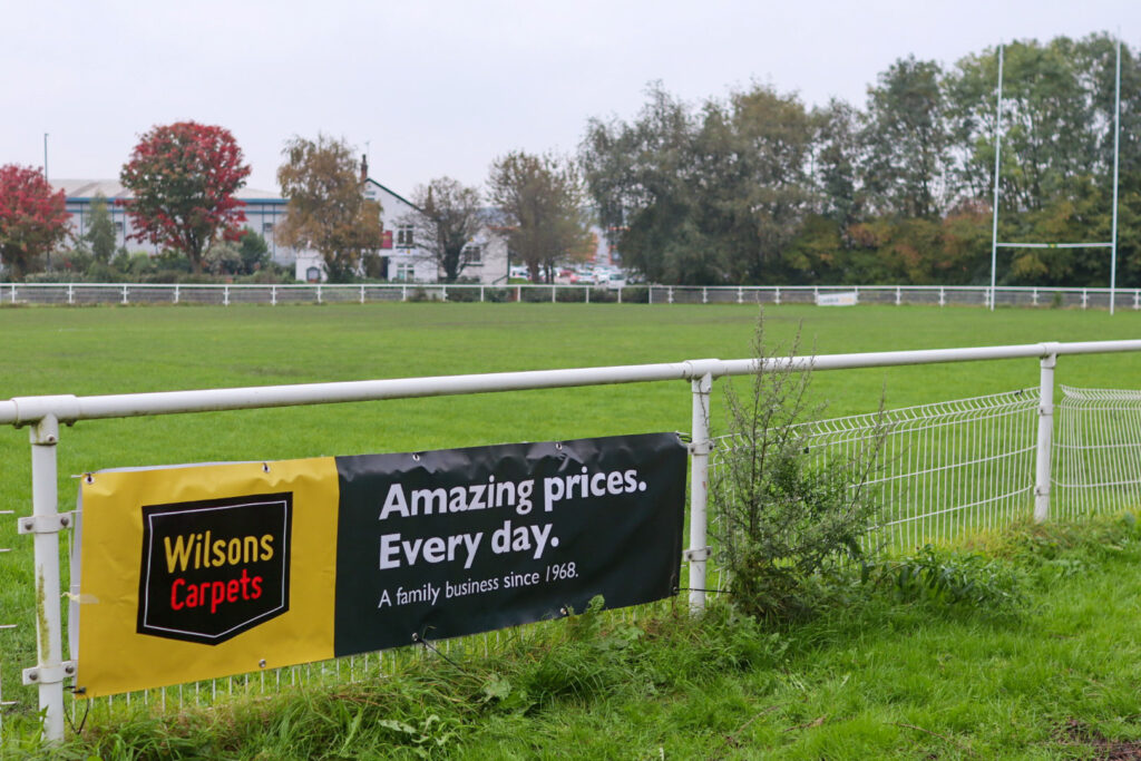 Pitch side advertising - Wilsons Carpets - Amazing Prices. Every day. A family business since 1968. A banner on the fence around a pitch. 
